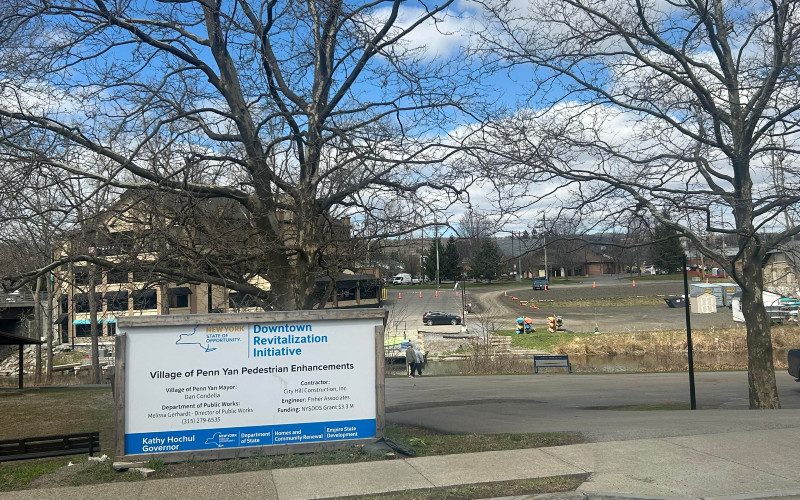 Sign for Penn Yan's Downtown Revitalization Initiative in front of Lake Street Park construction site, showing early progress with equipment and orange cones, leafless trees, and buildings in the background under a partly cloudy sky.